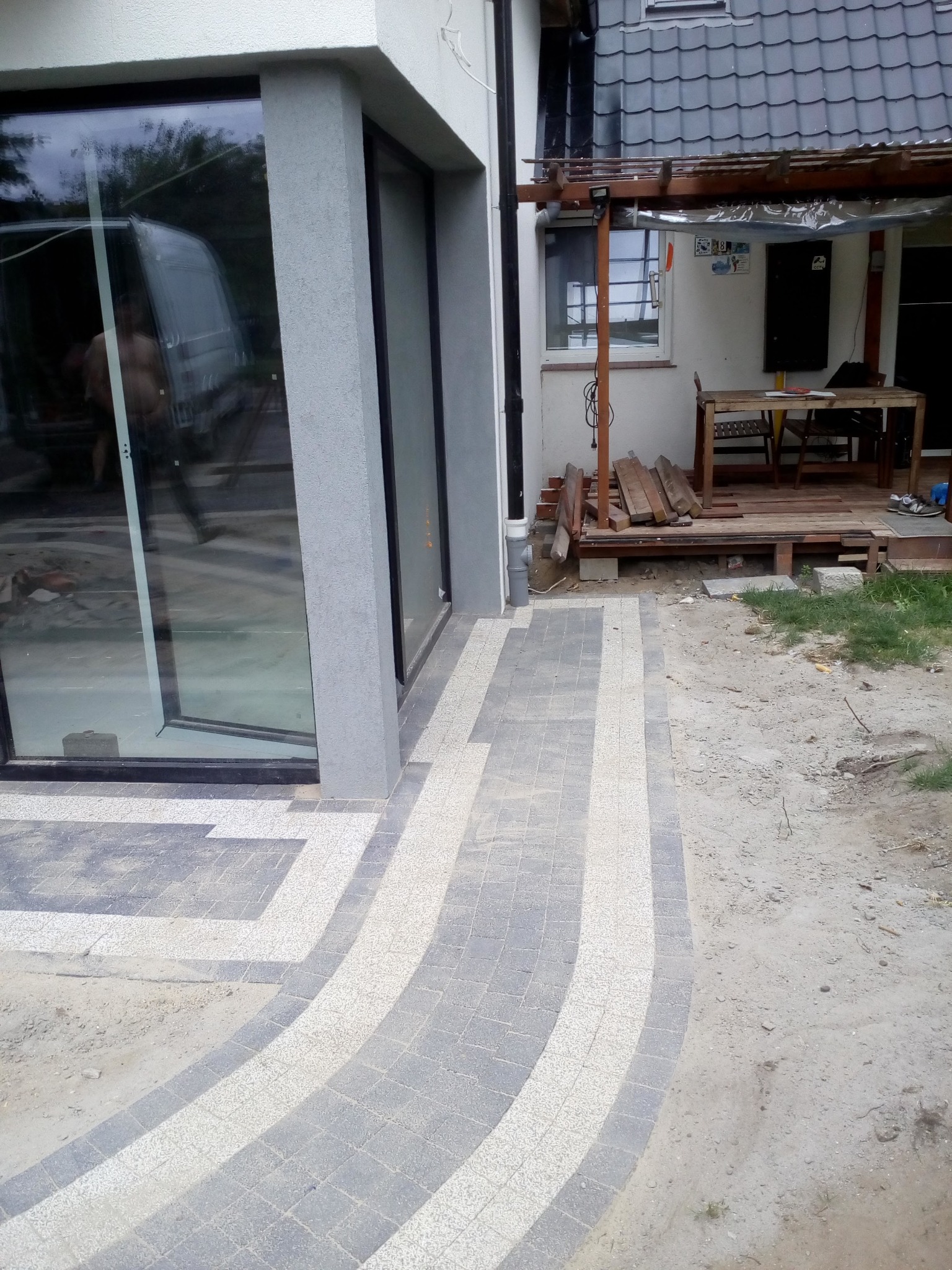 Ułożona kostka brukowa w odcieniach szarości i bieli tworząca wzór ścieżki przed nowoczesnym budynkiem z dużymi oknami, widoczne elementy tarasu i przygotowane deski.