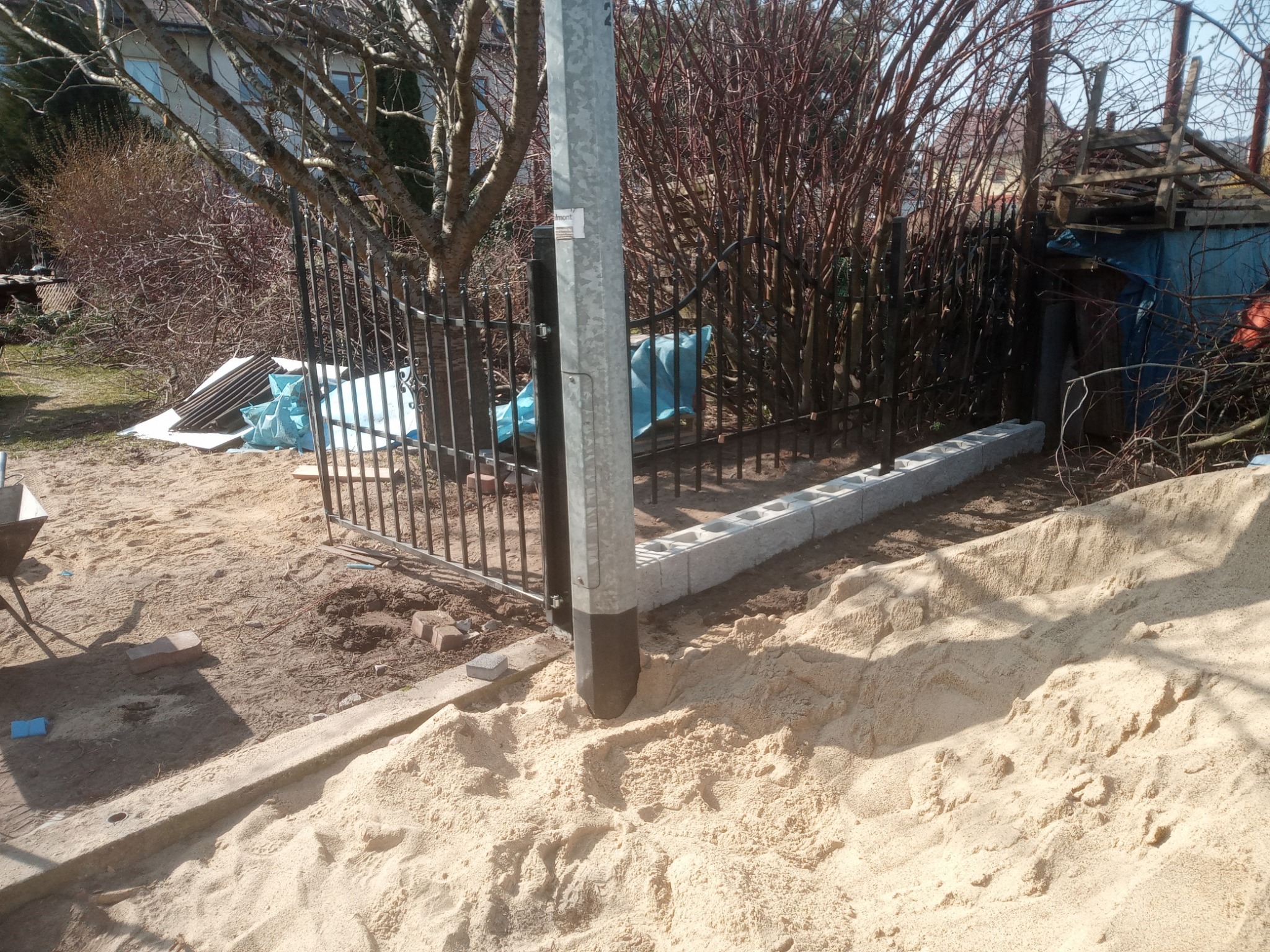 Przygotowanie podłoża z piasku pod montaż metalowego ogrodzenia z podmurówką z bloczków betonowych, widoczne narzędzia i materiały budowlane.