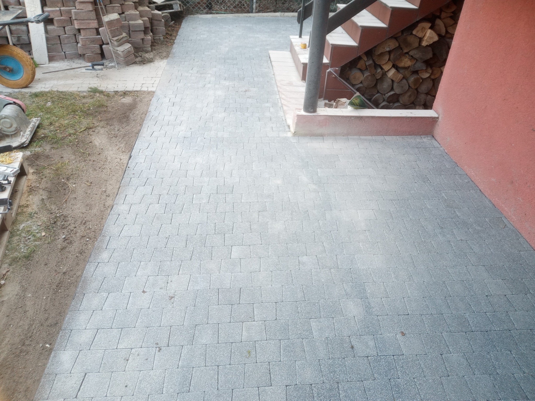 Świeżo ułożona, szara kostka brukowa prowadząca od trawnika do schodów z zapasem drewna pod nimi, widoczna ubijarka i stos kostki.