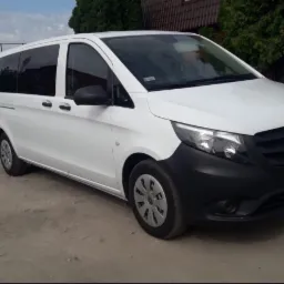 Biały Mercedes Vito, zaparkowany na szutrowej drodze, z widoczną tablicą rejestracyjną i elementami otoczenia jak drzewa i budynki.