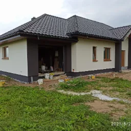 Jednorodzinny dom z ciemną dachówką i świeżo pomalowaną elewacją w kolorach beżu i brązu, widoczne wiaderka po farbie i pozostałości prac na trawniku.