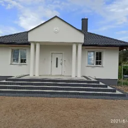 Nowoczesny dom jednorodzinny z białą elewacją, kolumnami przy wejściu i schodami z szarej kostki brukowej, widok z przodu, błękitne niebo z chmurami.