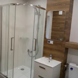 Nowoczesna łazienka z narożną kabiną prysznicową, białą umywalką z szafką i lustrem w drewnianej obudowie, widok na toaletę.