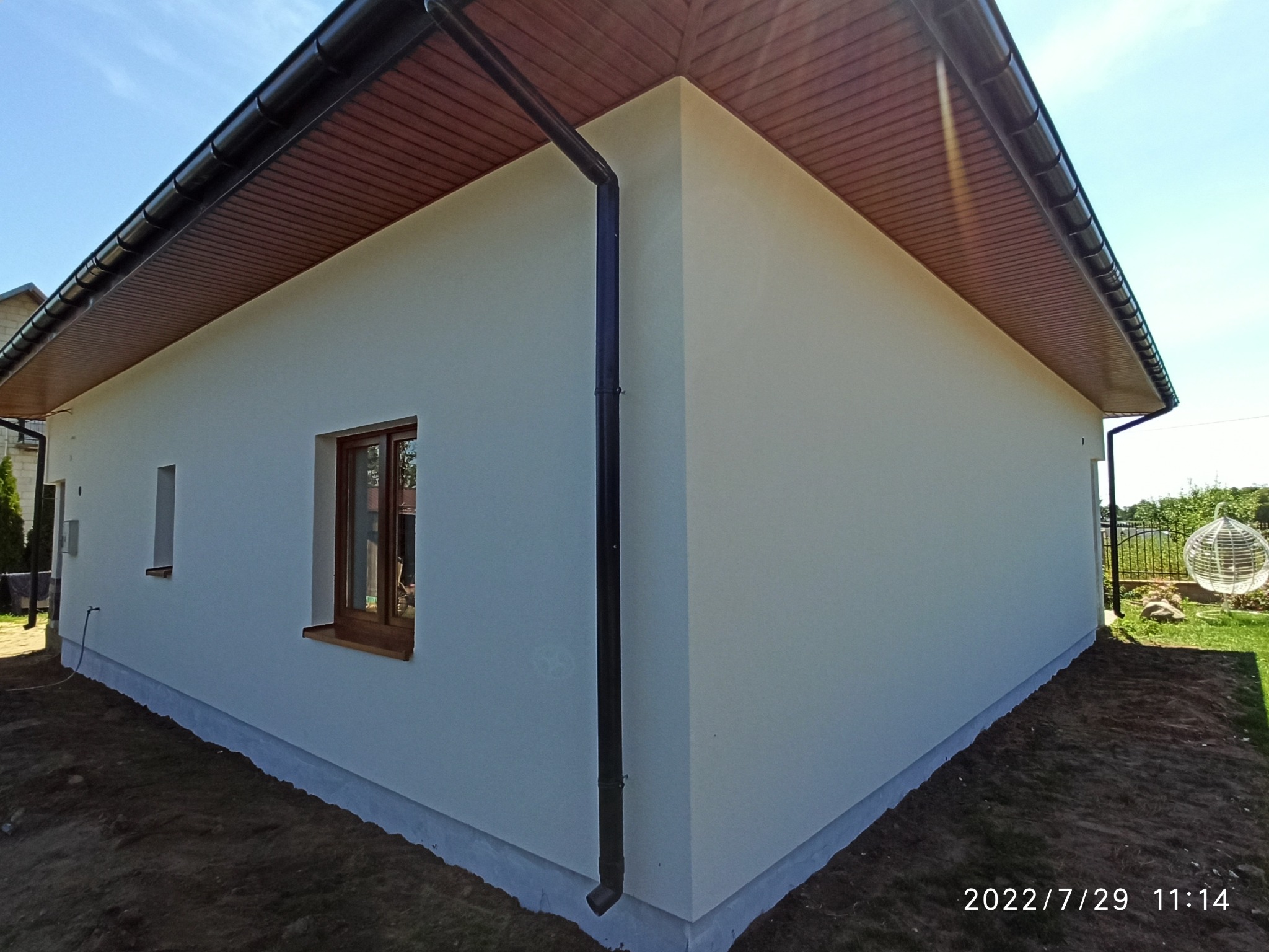 Widok z zewnątrz na jednopiętrowy dom z jasną elewacją, brązowymi drewnianymi oknami i ciemną rynną, z widocznym fragmentem ogrodu w tle.