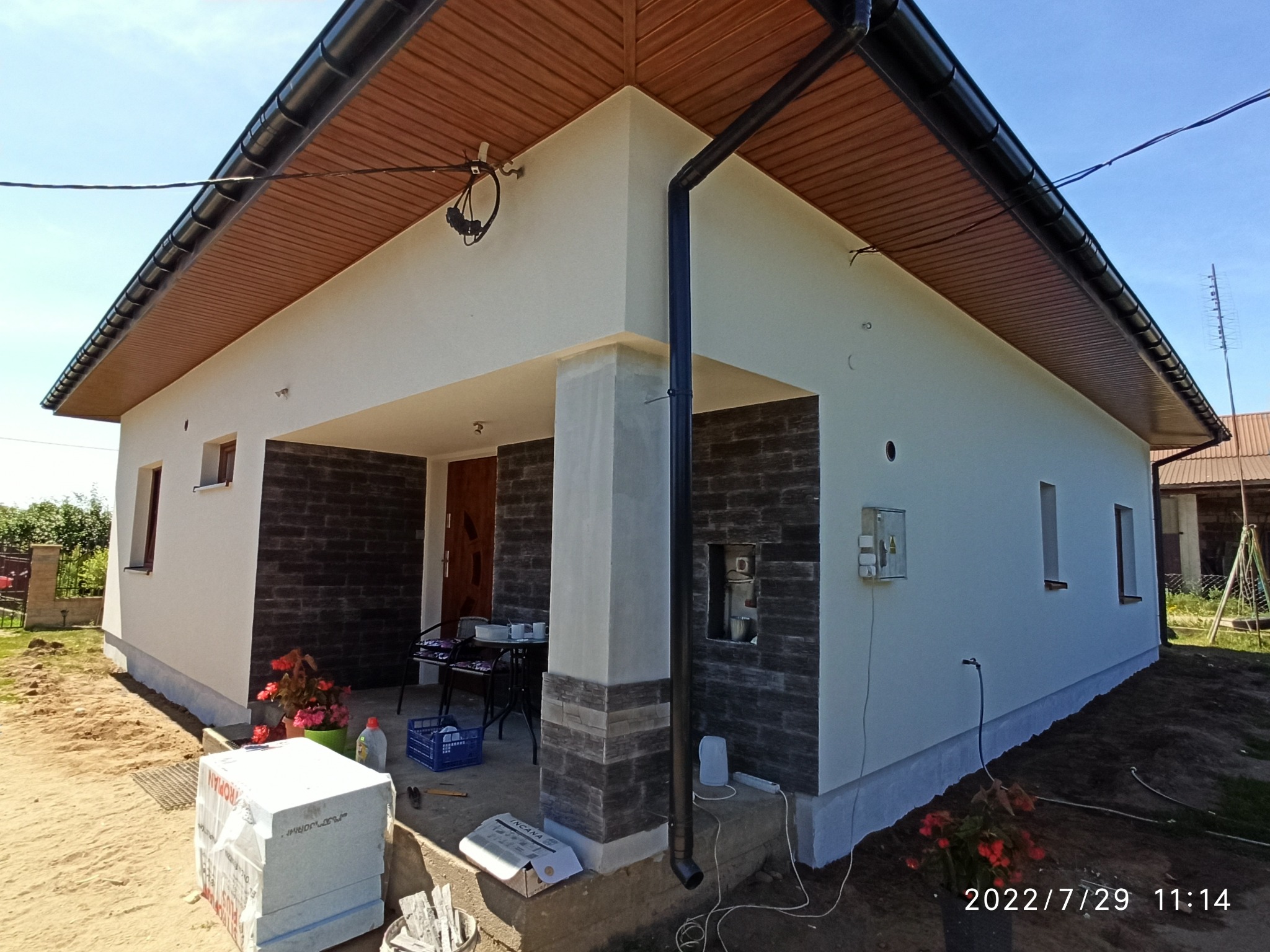 Parterowy dom jednorodzinny z jasną elewacją i fragmentami wykończonymi ciemną okładziną imitującą kamień, widoczny fragment ocieplenia styropianem na ziemi, rynna spustowa i elementy wykończeniowe...