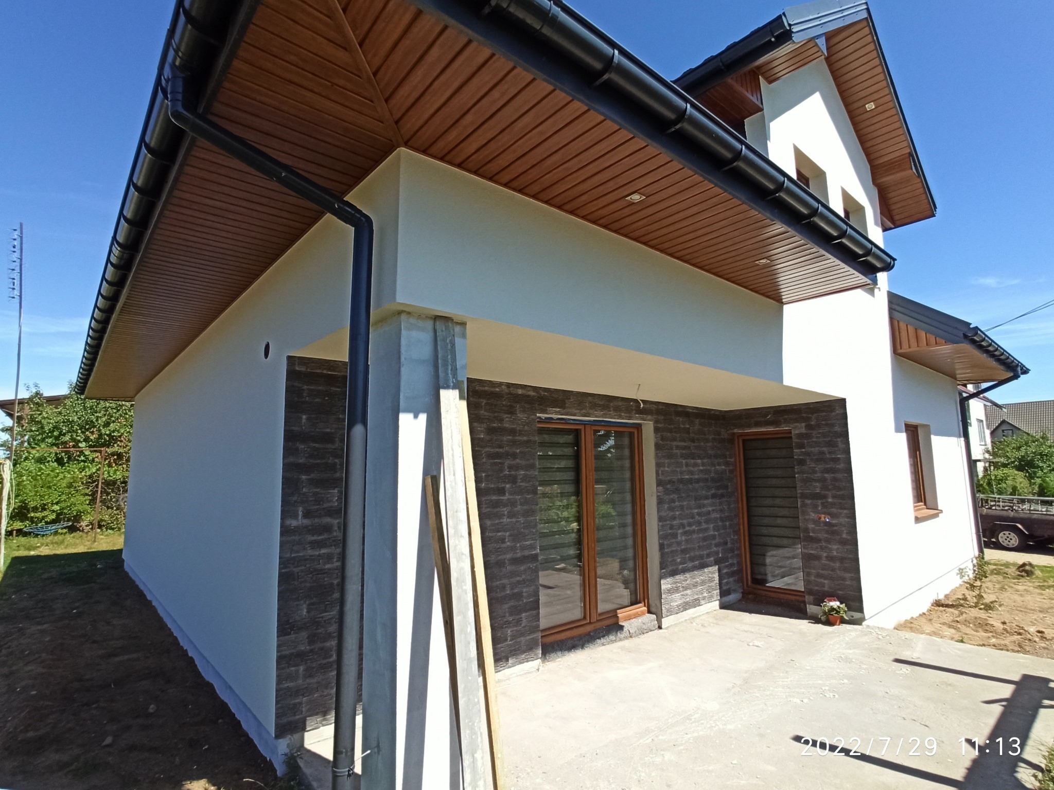 Nowoczesny dom jednorodzinny z jasnoszarą, gładką elewacją i fragmentem ściany wykończonym ciemnoszarą imitacją kamienia, widoczne pionowe deski opierające się o filar.