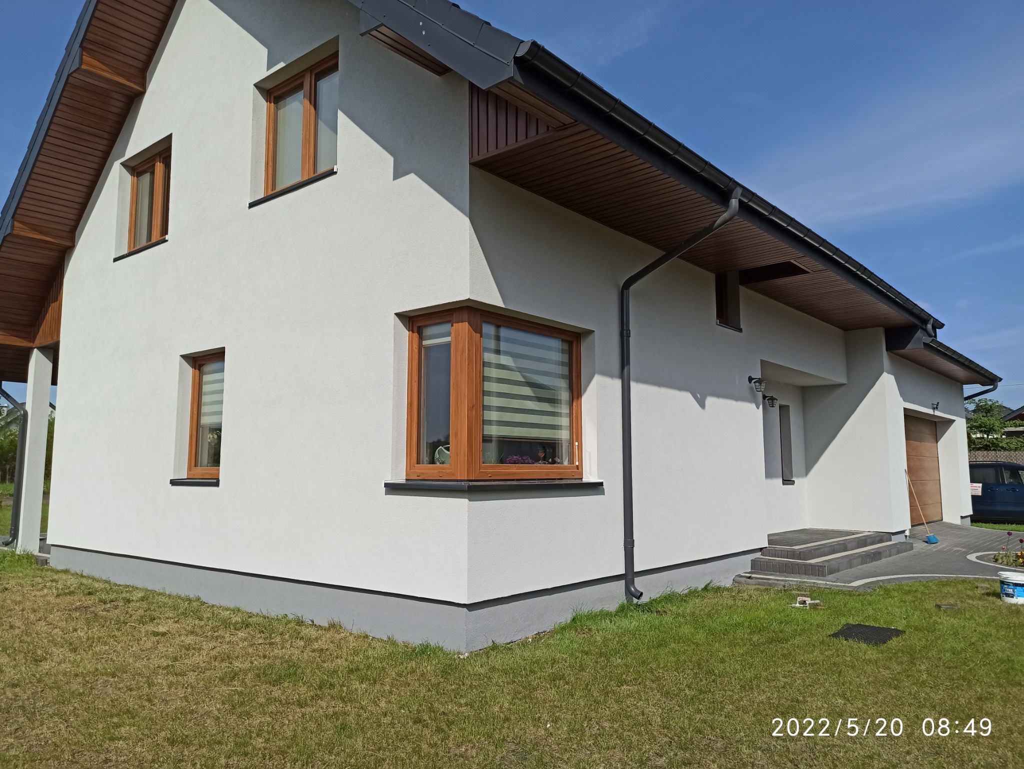 Świeżo otynkowana elewacja domu jednorodzinnego w kolorze białym, z drewnianymi oknami i szarą podmurówką, widoczna z boku na tle błękitnego nieba.
