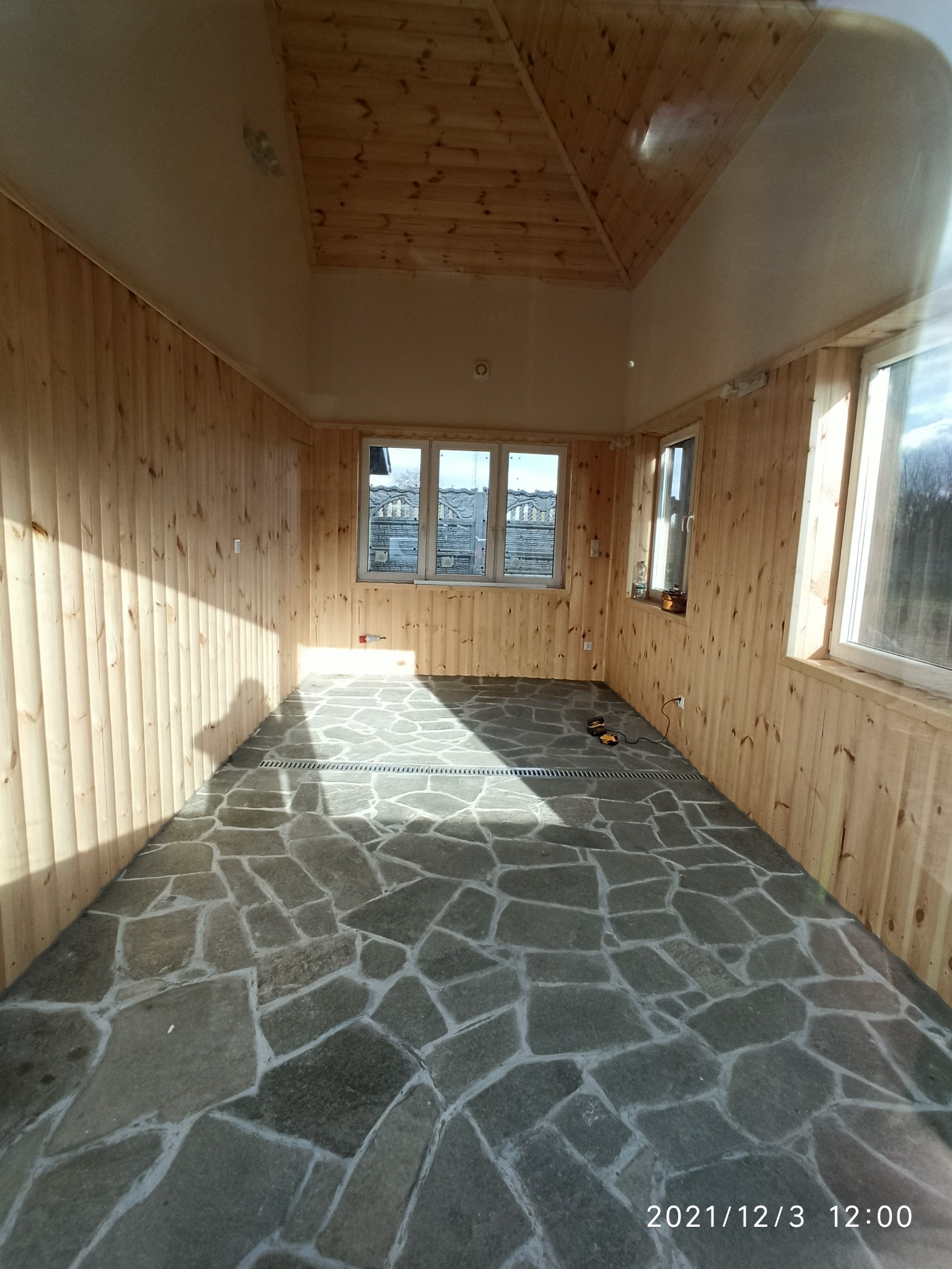 Wnętrze wykończonego pomieszczenia z drewnianą boazerią, kamienną podłogą z odpływem liniowym i oknami z widokiem na zewnątrz.