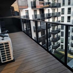 Maxiclean - Nowoczesny balkon z drewnianą podłogą, szklaną balustradą i widokiem na inne balkony apartamentowca; na podłodze stoi biały klimatyzator z dwiema dekoracyjnymi figurkami ptaków.