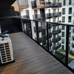 Nowoczesny balkon z drewnianą podłogą, szklaną balustradą i widokiem na inne balkony apartamentowca; na podłodze stoi biały klimatyzator z dwiema dekoracyjnymi figurkami ptaków.
