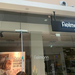 Maxiclean - Osoba myje okno wystawowe sklepu optycznego Fielmann za pomocą mopa z mikrofibry na długim kiju, widoczne plakaty reklamowe i logo sklepu.