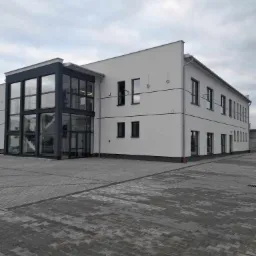 Nowoczesny, dwukondygnacyjny budynek biurowy z przeszklonym wejściem, widoczny z poziomu brukowanego placu w pochmurny dzień.