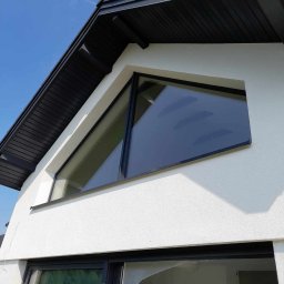 Maxiclean - Nowoczesny dom z białymi ścianami i czarną dachówką, z nietypowym trójkątnym oknem i dużymi przesuwnymi drzwiami balkonowymi zabezpieczonymi folią.