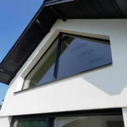 Nowoczesny dom z białymi ścianami i czarną dachówką, z nietypowym trójkątnym oknem i dużymi przesuwnymi drzwiami balkonowymi zabezpieczonymi folią.