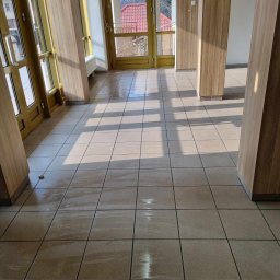 Maxiclean - Wnętrze budynku z wypolerowaną, mokrą podłogą z płytek, drewnianymi panelami i drzwiami wejściowymi przepuszczającymi światło słoneczne.