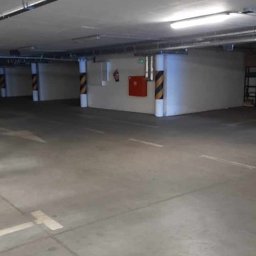 Maxiclean - Pusta przestrzeń garażowa z betonową podłogą, oznaczonymi miejscami parkingowymi, oświetleniem sufitowym i kolumnami w czarno-żółte pasy.