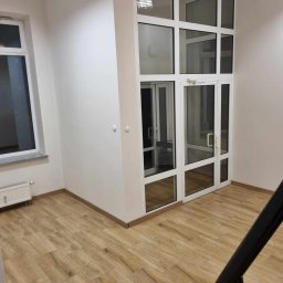 Maxiclean - Wnętrze biura z drewnopodobną podłogą i przeszklonym wejściem z białymi ramami, widoczne schody z czarną barierką.