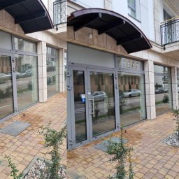 Maxiclean - Fasada budynku biurowego z dużymi, szklanymi oknami i drzwiami wejściowymi, widoczna z zewnątrz na tle ulicy i chodnika wyłożonego kostką brukową.