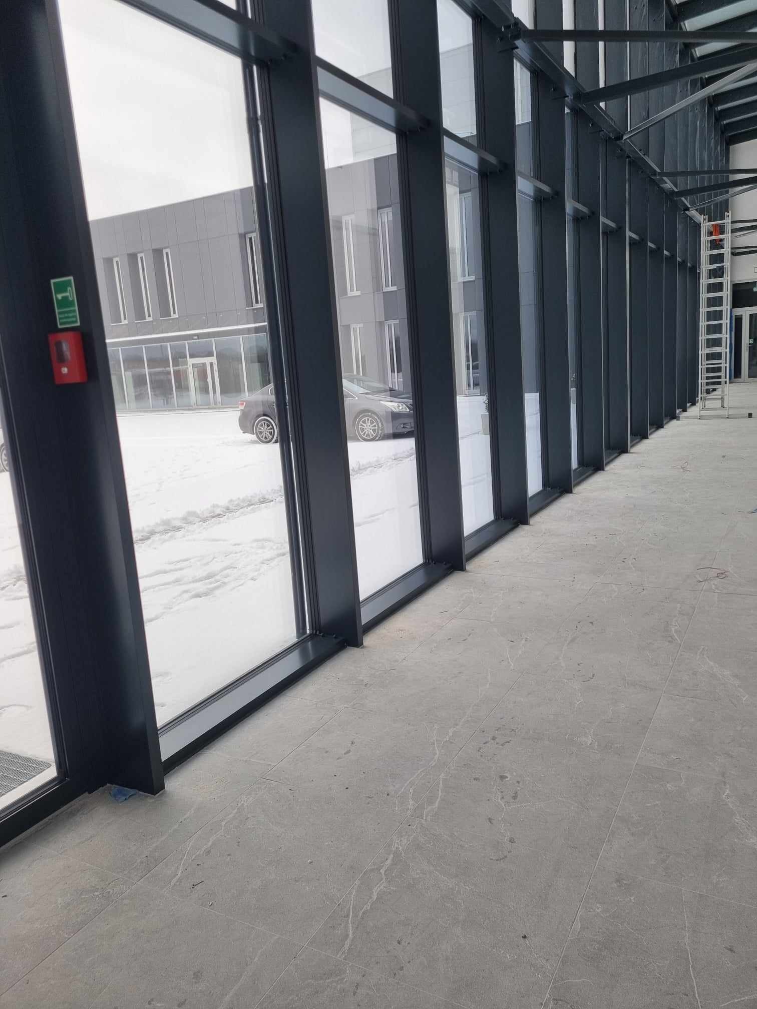 Wnętrze budynku z widokiem na zaśnieżony parking przez duże, regularnie rozmieszczone okna z czarnymi ramami. W tle budynek biurowy i metalowa drabina.