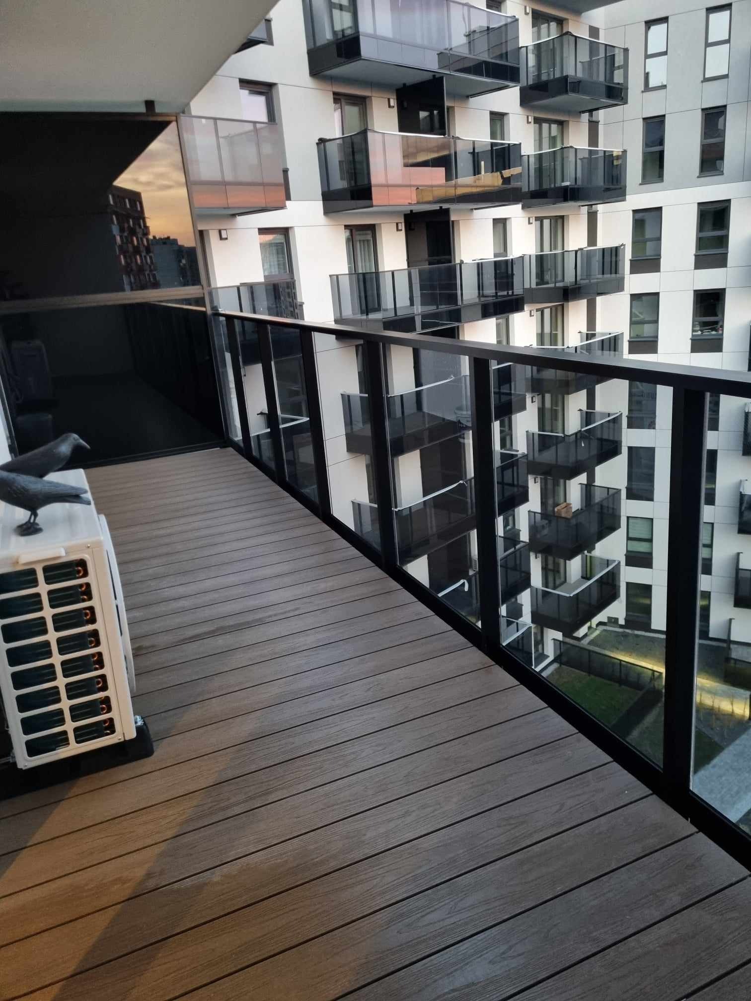 Nowoczesny balkon z drewnianą podłogą, szklaną balustradą i widokiem na inne balkony apartamentowca; na podłodze stoi biały klimatyzator z dwiema dekoracyjnymi figurkami ptaków.