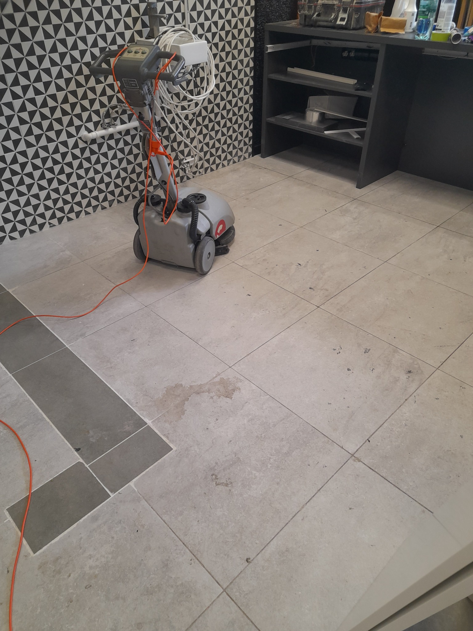 Szorowarka do podłóg na kafelkach w trakcie pracy, widoczne plamy i zabrudzenia, tło stanowią czarno-białe trójkątne płytki na ścianie i półki z wyposażeniem.