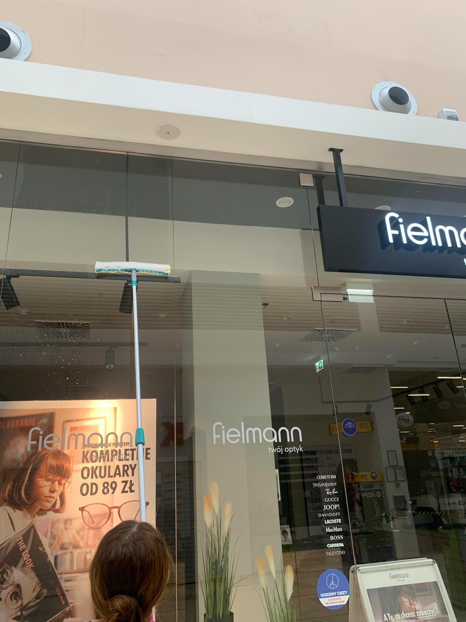 Osoba myje okno wystawowe sklepu optycznego Fielmann za pomocą mopa z mikrofibry na długim kiju, widoczne plakaty reklamowe i logo sklepu.