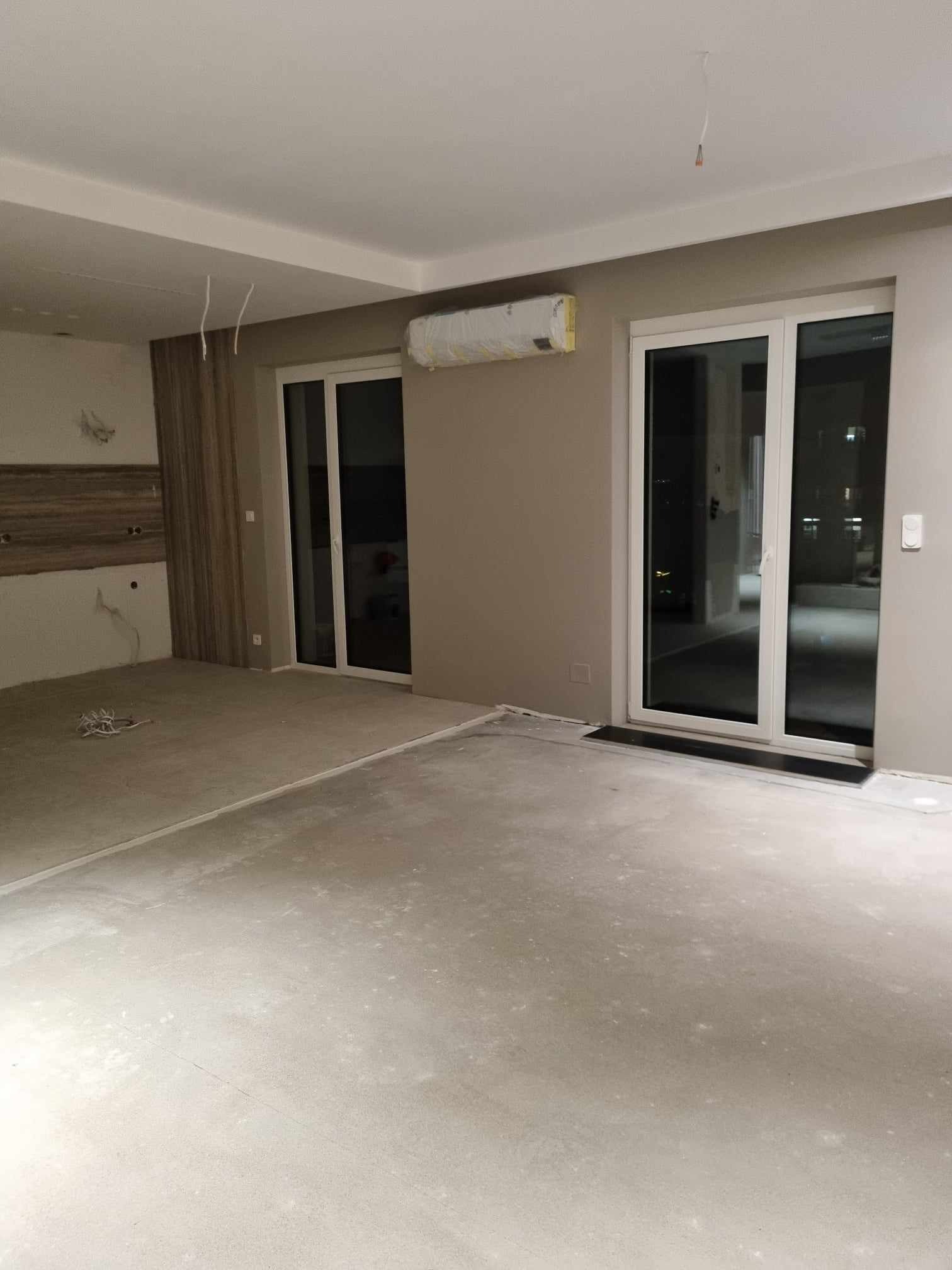 Surowe wnętrze mieszkania w trakcie remontu, widoczne betonowe podłoże, niezabezpieczony klimatyzator, duże przesuwne okna balkonowe i wystające kable ze stropu.