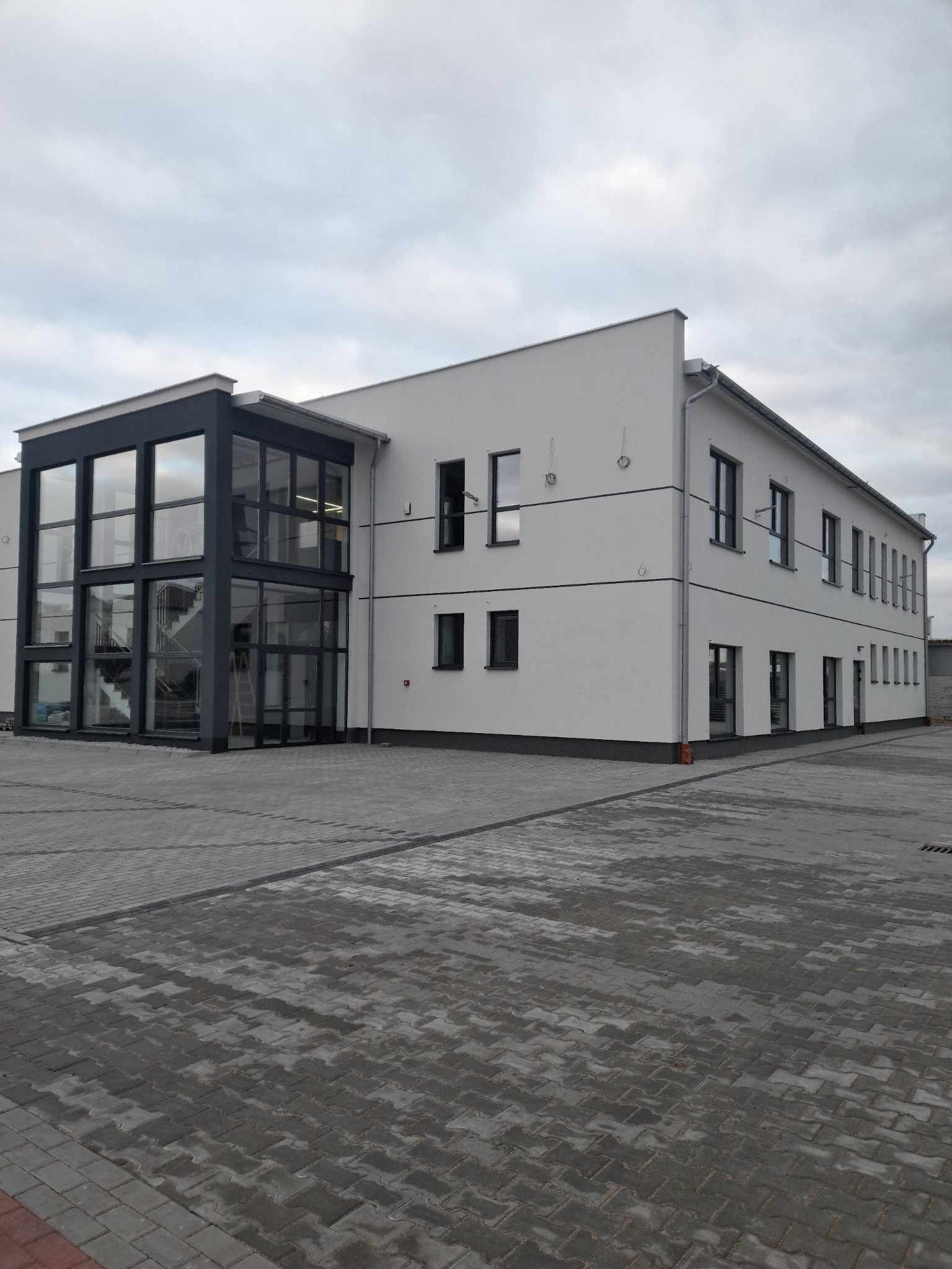 Nowoczesny, dwukondygnacyjny budynek biurowy z przeszklonym wejściem, widoczny z poziomu brukowanego placu w pochmurny dzień.