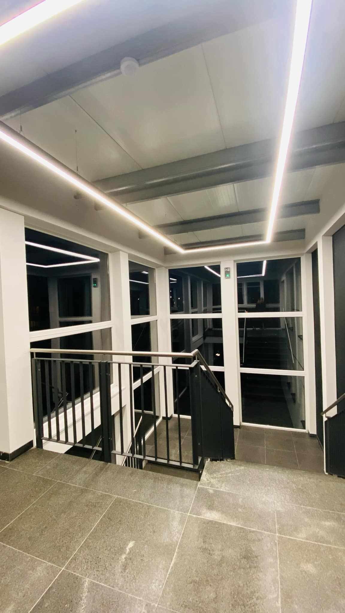 Wnętrze klatki schodowej z szarymi płytkami podłogowymi, czarnymi metalowymi barierkami i białymi ścianami, oświetlone liniowymi lampami LED na suficie.