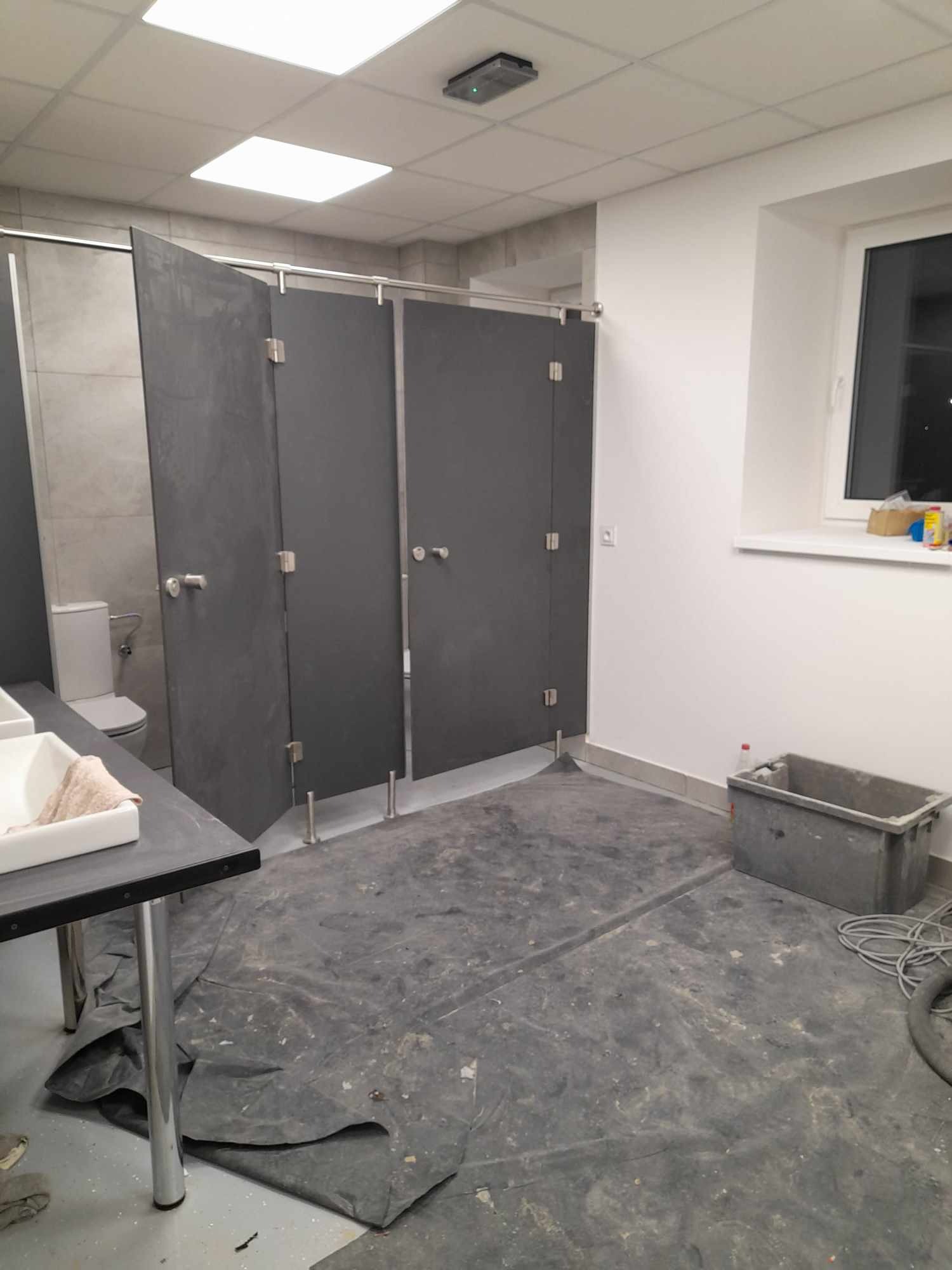 Wnętrze publicznej toalety w trakcie sprzątania po remoncie: widoczne kabiny WC z szarego laminatu, zabezpieczona folią podłoga i pojemnik na odpady budowlane.
