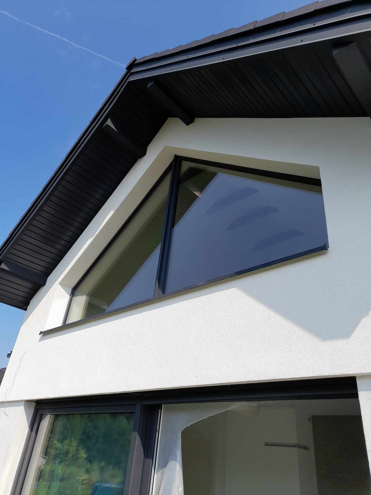 Nowoczesny dom z białymi ścianami i czarną dachówką, z nietypowym trójkątnym oknem i dużymi przesuwnymi drzwiami balkonowymi zabezpieczonymi folią.