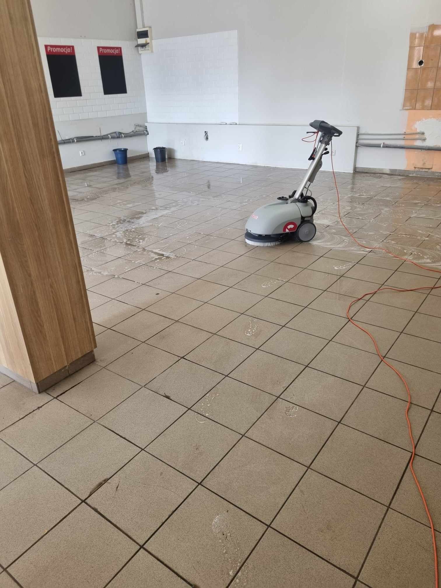 Profesjonalna maszyna do mycia podłóg w trakcie pracy na kafelkach w dużym pomieszczeniu, widoczne ślady wody i piany, w tle ściany z elementami instalacji i tablicami.