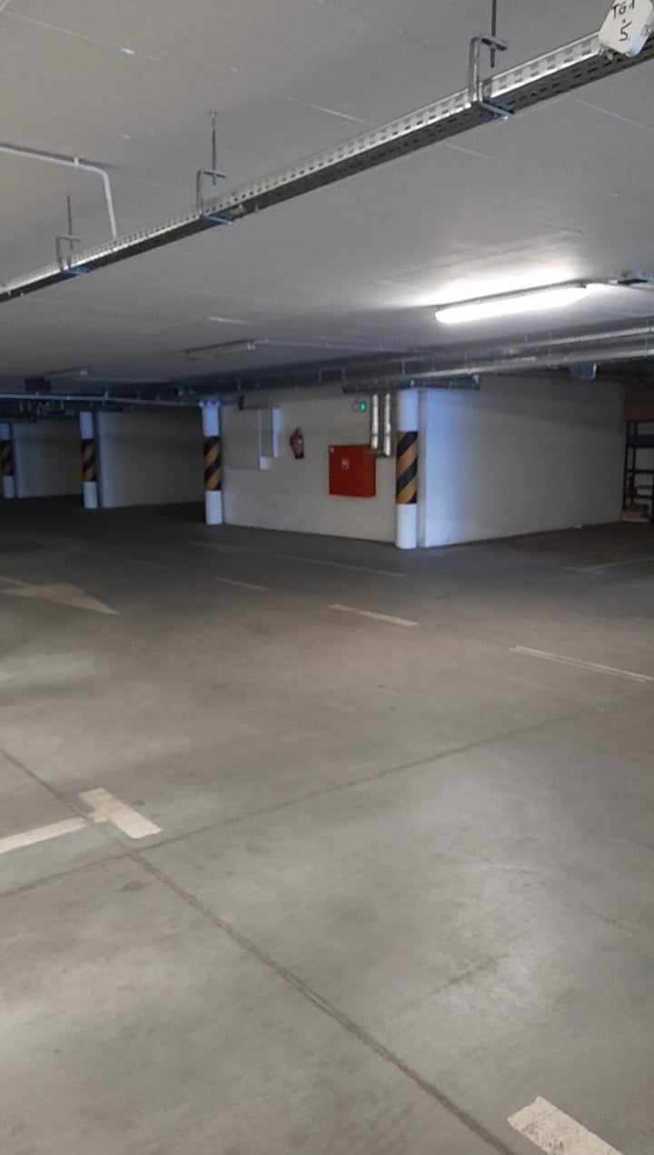 Pusta przestrzeń garażowa z betonową podłogą, oznaczonymi miejscami parkingowymi, oświetleniem sufitowym i kolumnami w czarno-żółte pasy.