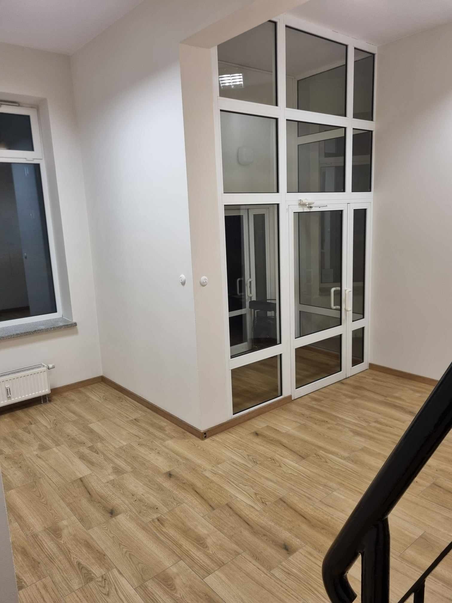 Wnętrze biura z drewnopodobną podłogą i przeszklonym wejściem z białymi ramami, widoczne schody z czarną barierką.