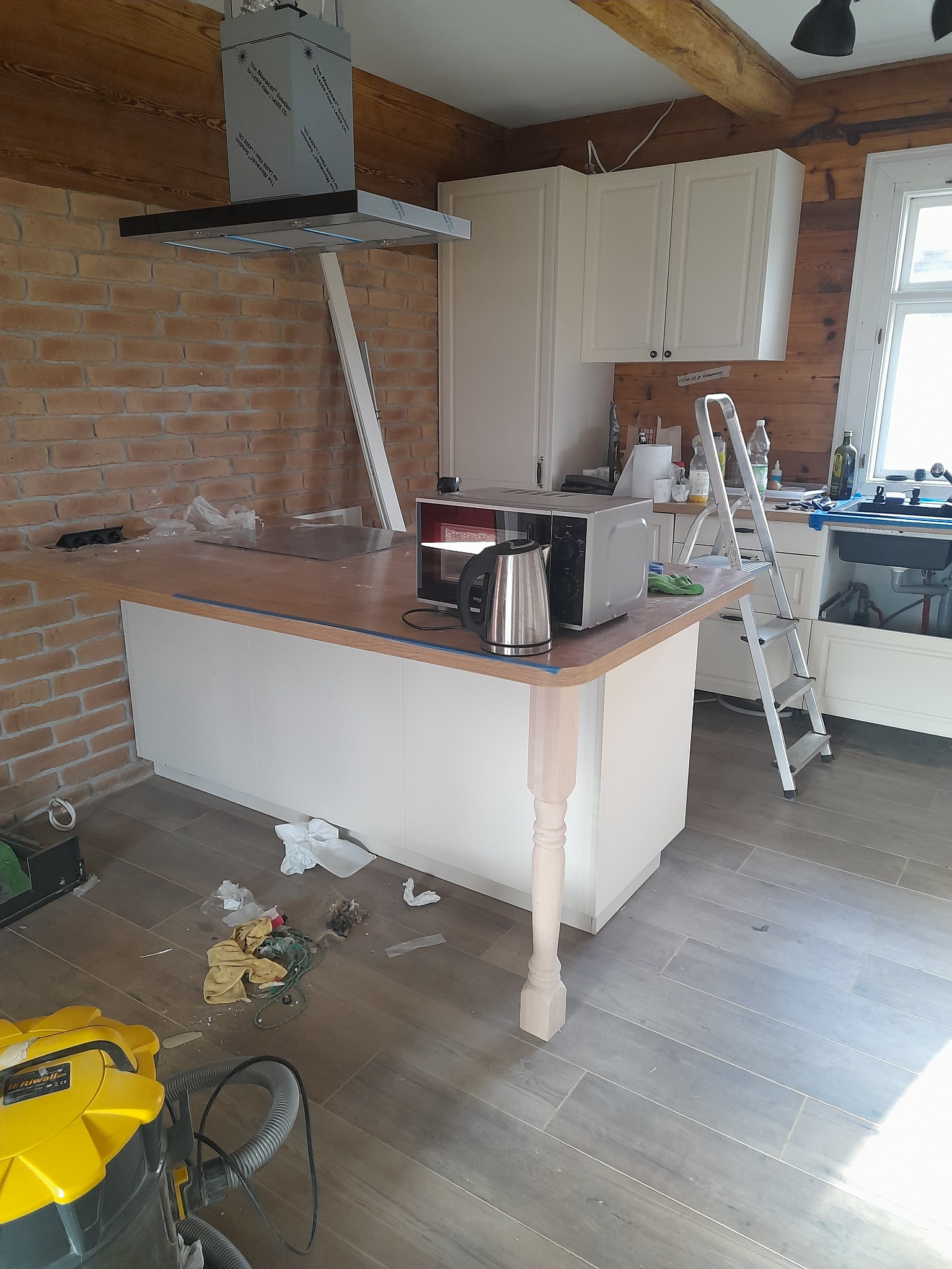 Niedokończona kuchnia z ceglaną ścianą, wyspą z niedomalowaną nogą, żółtym odkurzaczem przemysłowym i resztkami po remoncie na podłodze.