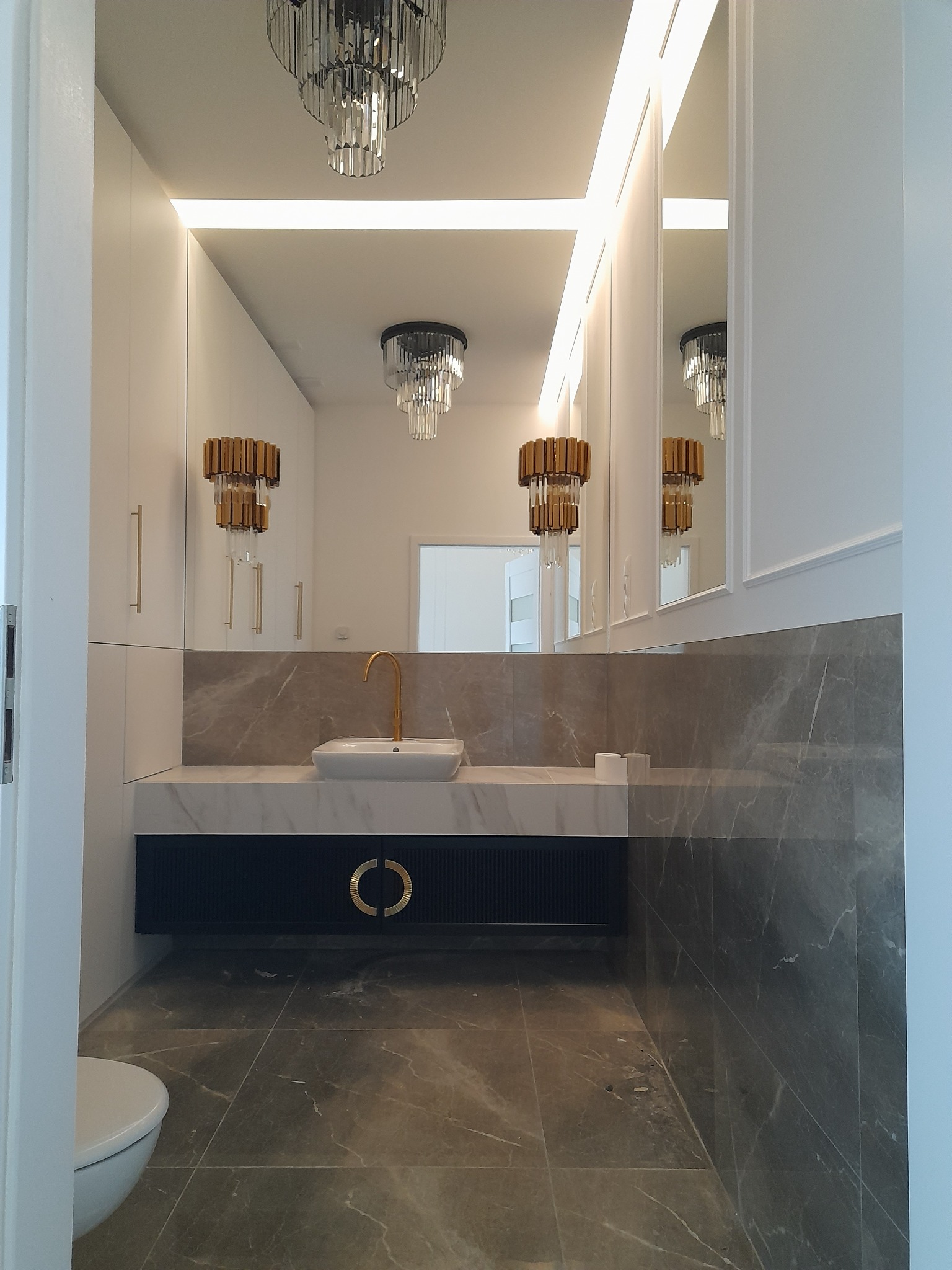 Elegancka, nowocześnie urządzona łazienka w Wasilkowie z marmurowymi płytkami na podłodze i ścianie, białą umywalką z pozłacaną armaturą oraz szafką z dekoracyjnymi, okrągłymi uchwytami. W tle...