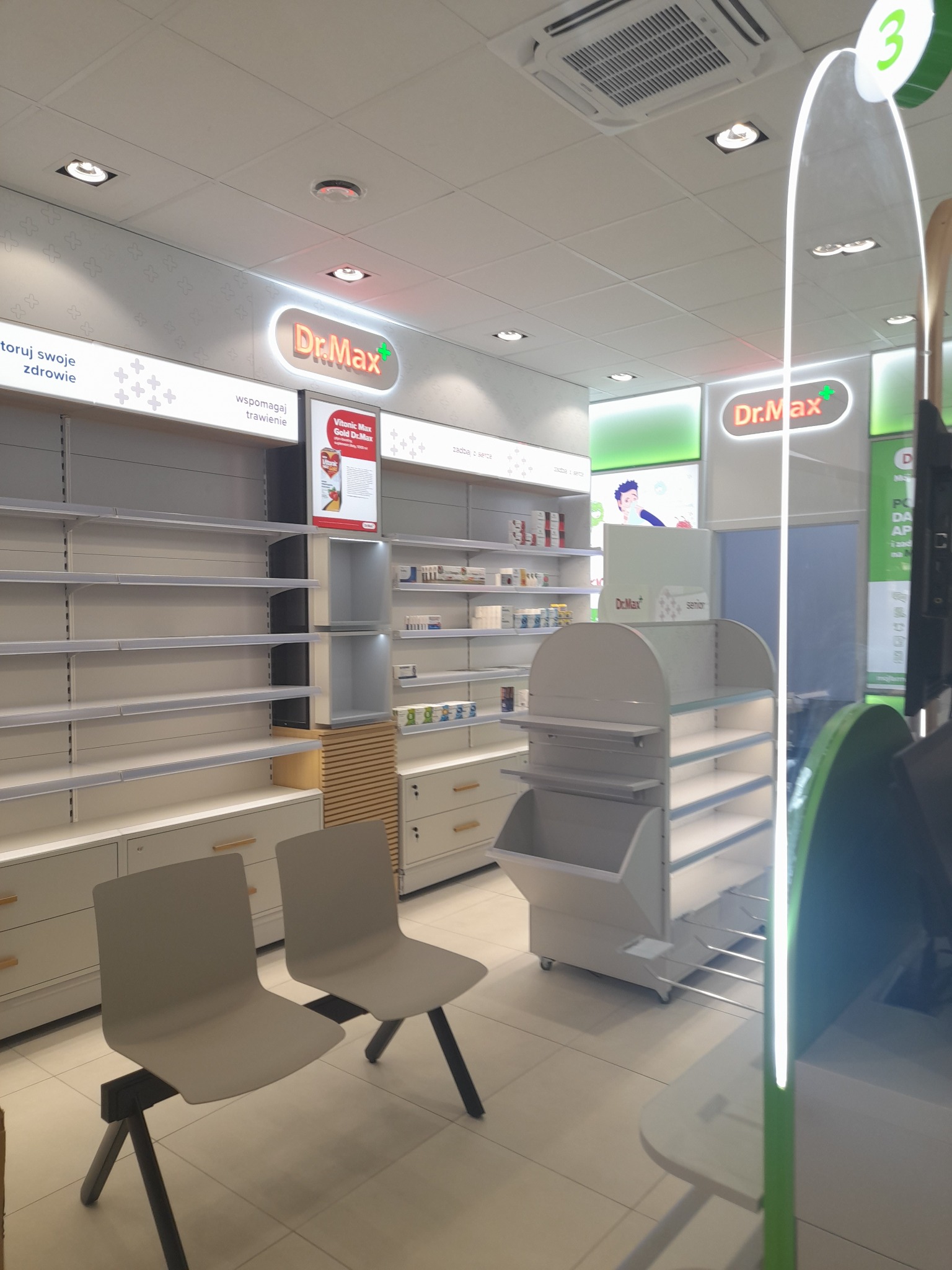 Wnętrze apteki sieci Dr.Max z pustymi półkami, dwoma krzesłami dla klientów i regałami ekspozycyjnymi w odcieniach bieli i szarości.