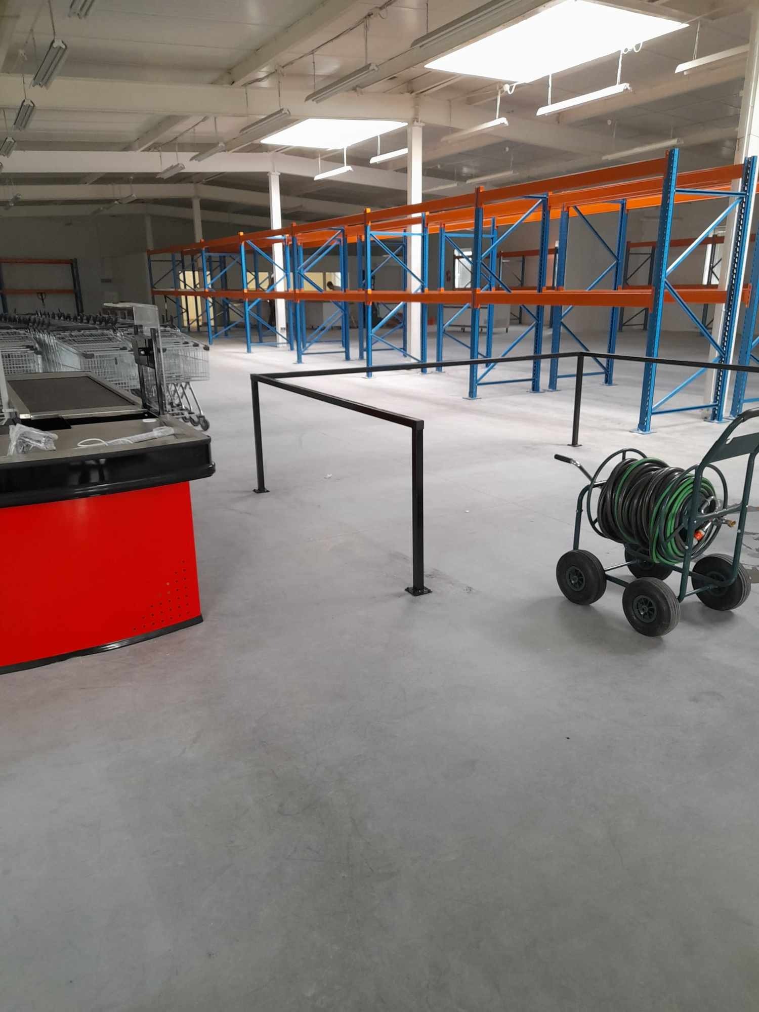 Wnętrze hali magazynowej lub sklepu w trakcie przygotowań: puste regały, wózki na zakupy, kasa w folii, wózek z wężem do mycia podłogi.