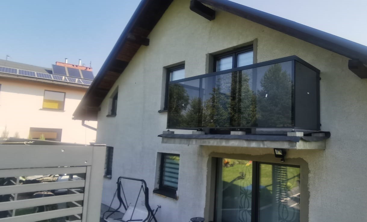 Balkon z czarną, szklaną balustradą na tle domu z beżową elewacją i widokiem na sąsiedni budynek z panelami słonecznymi na dachu.