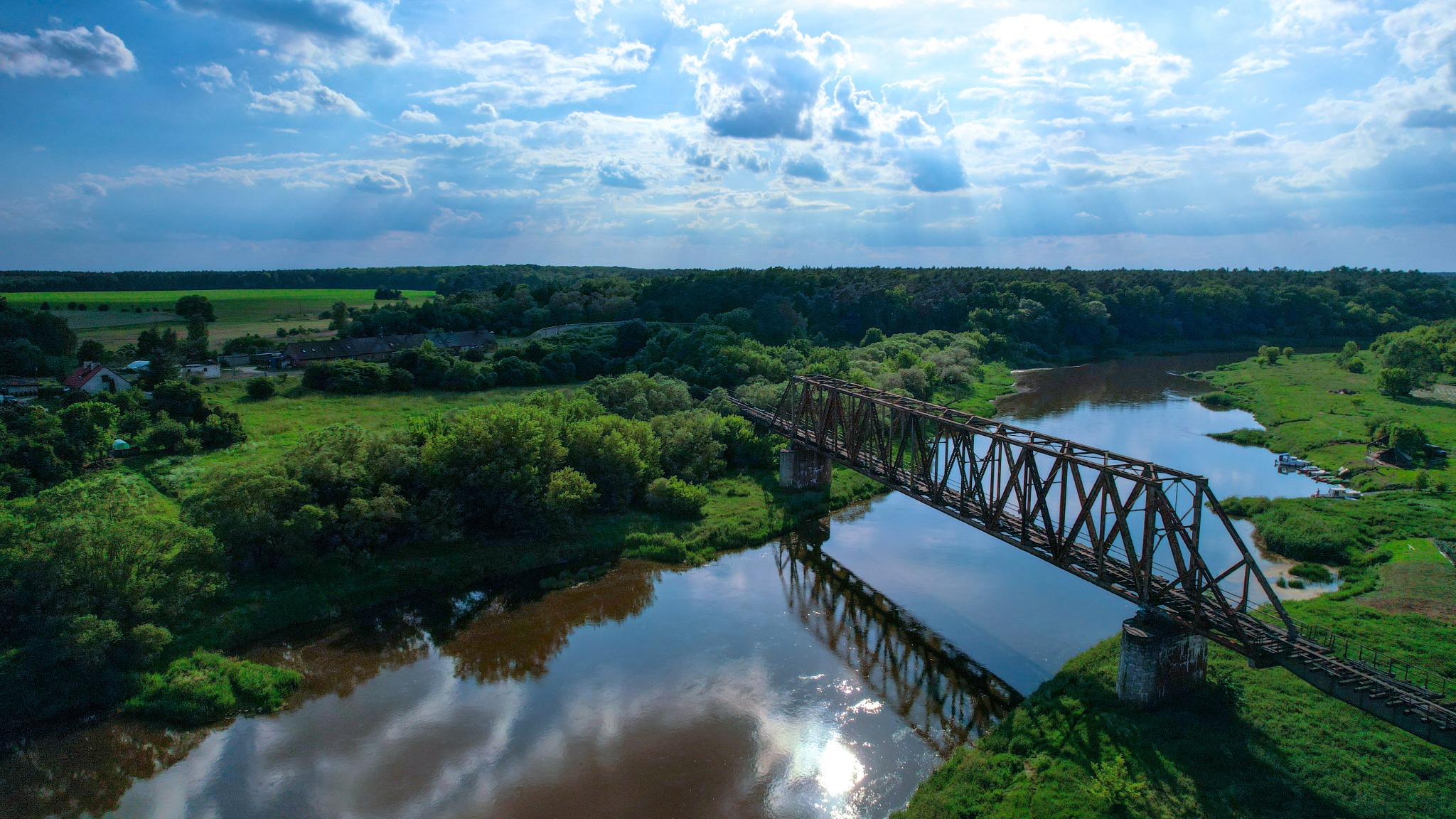 Szeroki kadr z drona przedstawiający stary, metalowy most kolejowy nad rzeką w okolicach Mrowina, otoczony bujną zielenią i lasami, z odbiciem chmur na powierzchni wody.