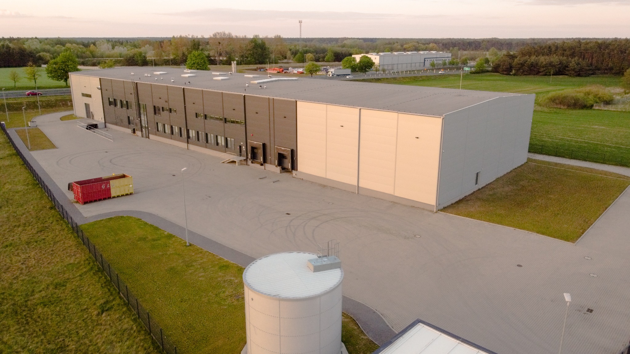 Zdjęcie z lotu ptaka przedstawia duży, nowoczesny budynek magazynowy z rampami załadunkowymi, otoczony zielenią i parkingiem. W tle widoczny las i inne budynki.