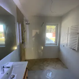 Nowoczesna łazienka z kabiną prysznicową typu walk-in, białą umywalką z szafką z drewnianymi akcentami, grzejnikiem drabinkowym i podłogą wyłożoną płytkami w jodełkę, widok z okna na zewnątrz.