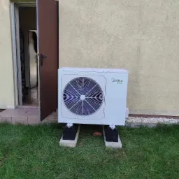 Biała jednostka zewnętrzna pompy ciepła Midea, ustawiona na betonowych bloczkach przed budynkiem z otwartymi drzwiami wejściowymi, widoczna trawa i białe kamienie.