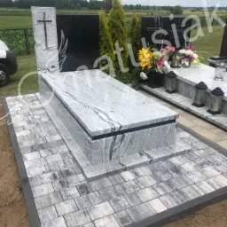 Jasnoszary nagrobek z granitu z krzyżem, otoczony szarą kostką brukową na cmentarzu, w tle zieleń.