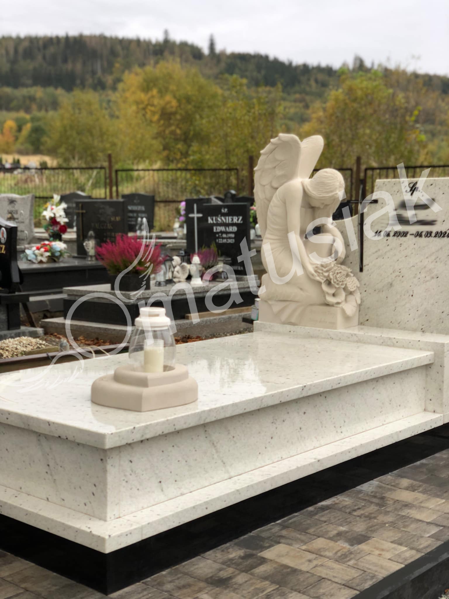 Jasny, granitowy pomnik z rzeźbą anioła, z widoczną datą na tablicy i lampionem na płycie, na tle cmentarza i zalesionego wzgórza.