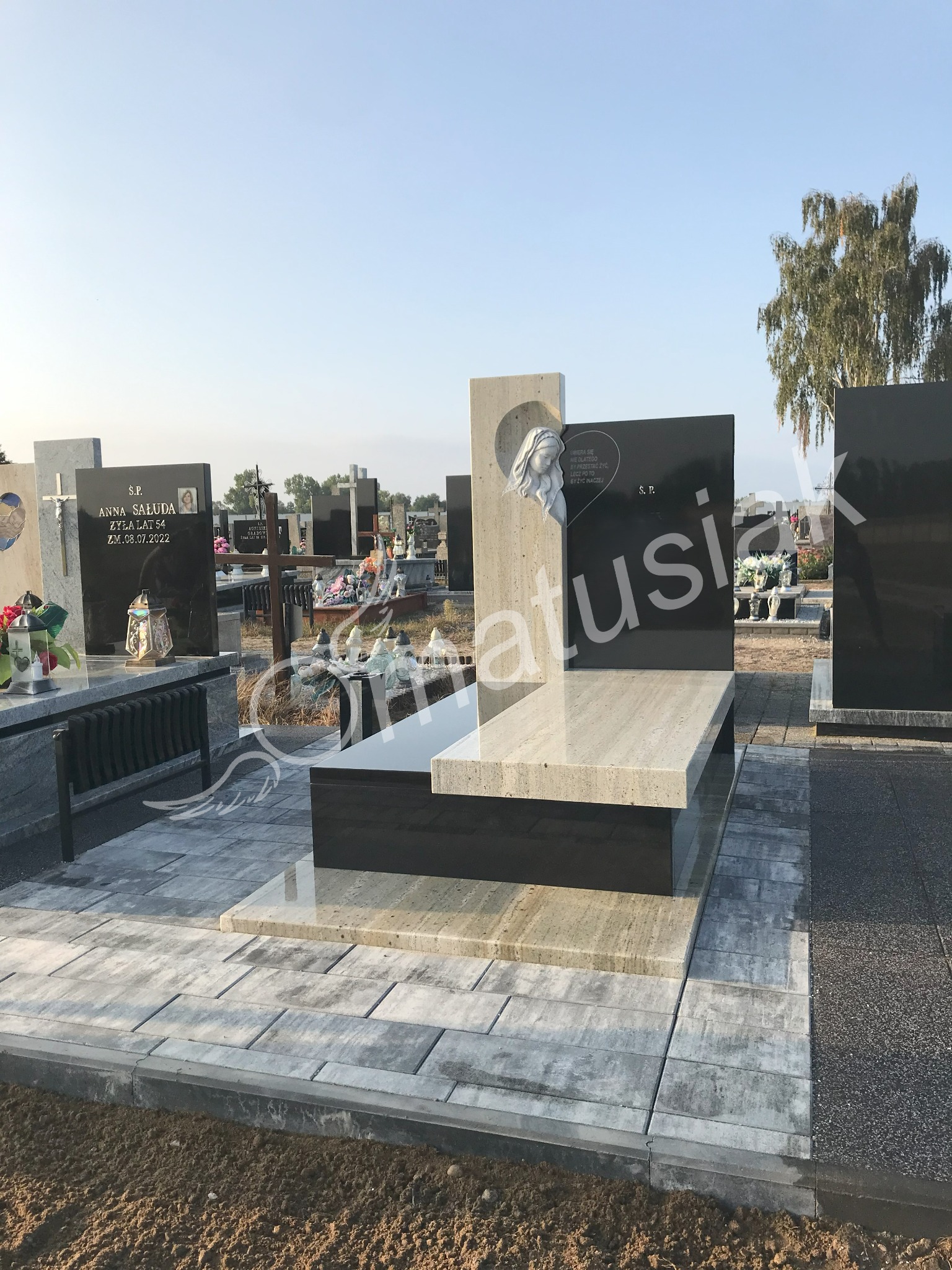 Nowoczesny, dwukolorowy pomnik z jasnego i czarnego granitu na cmentarzu, z rzeźbą kobiety wkomponowaną w jasny element. Widoczne inne groby w tle.