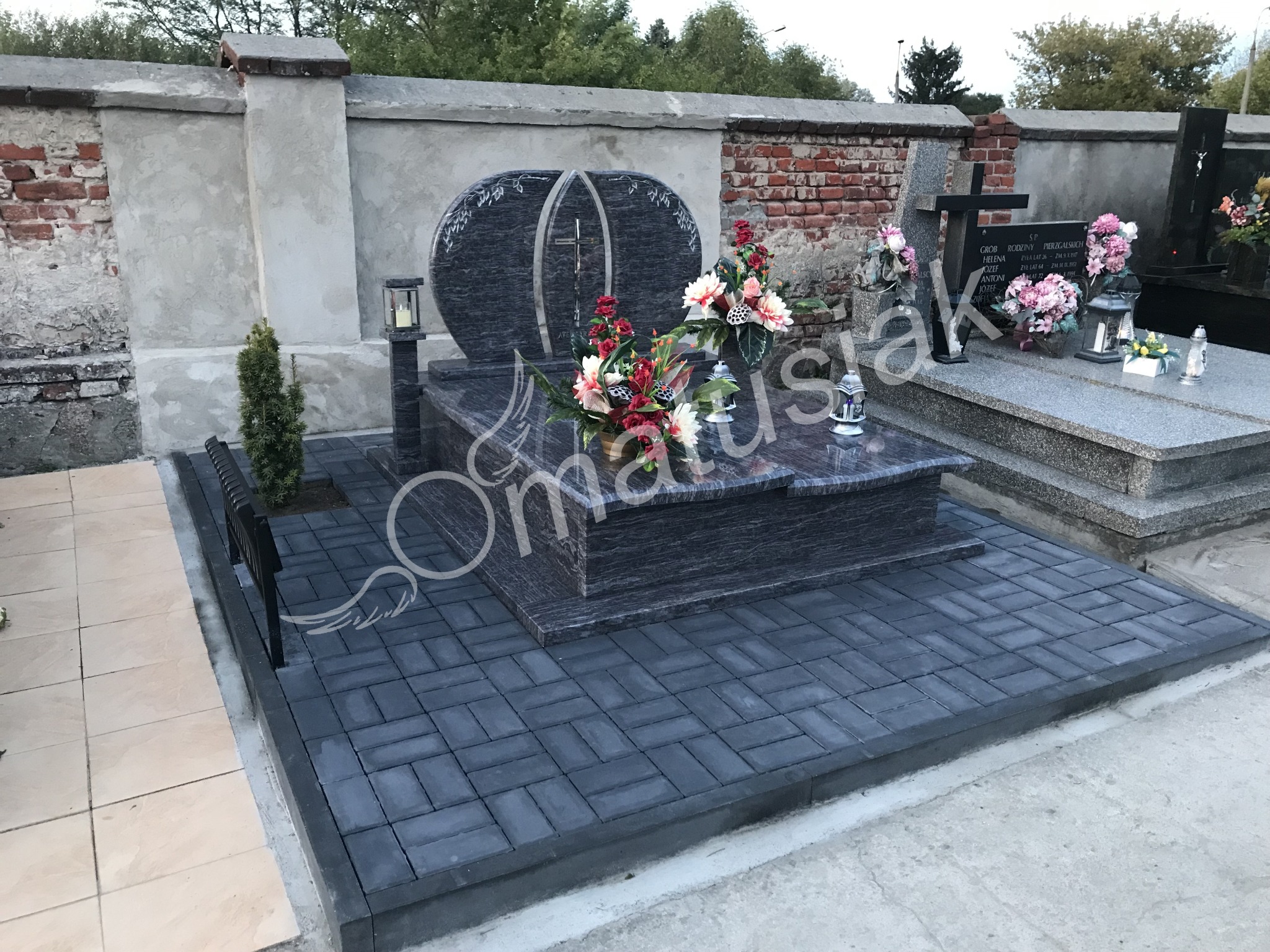 Podwójny, grafitowy pomnik z dwoma połączonymi owalami i krzyżem wewnątrz, otoczony kwiatami i lampionami, wyłożony ciemną kostką brukową, z małą ławeczką z boku.