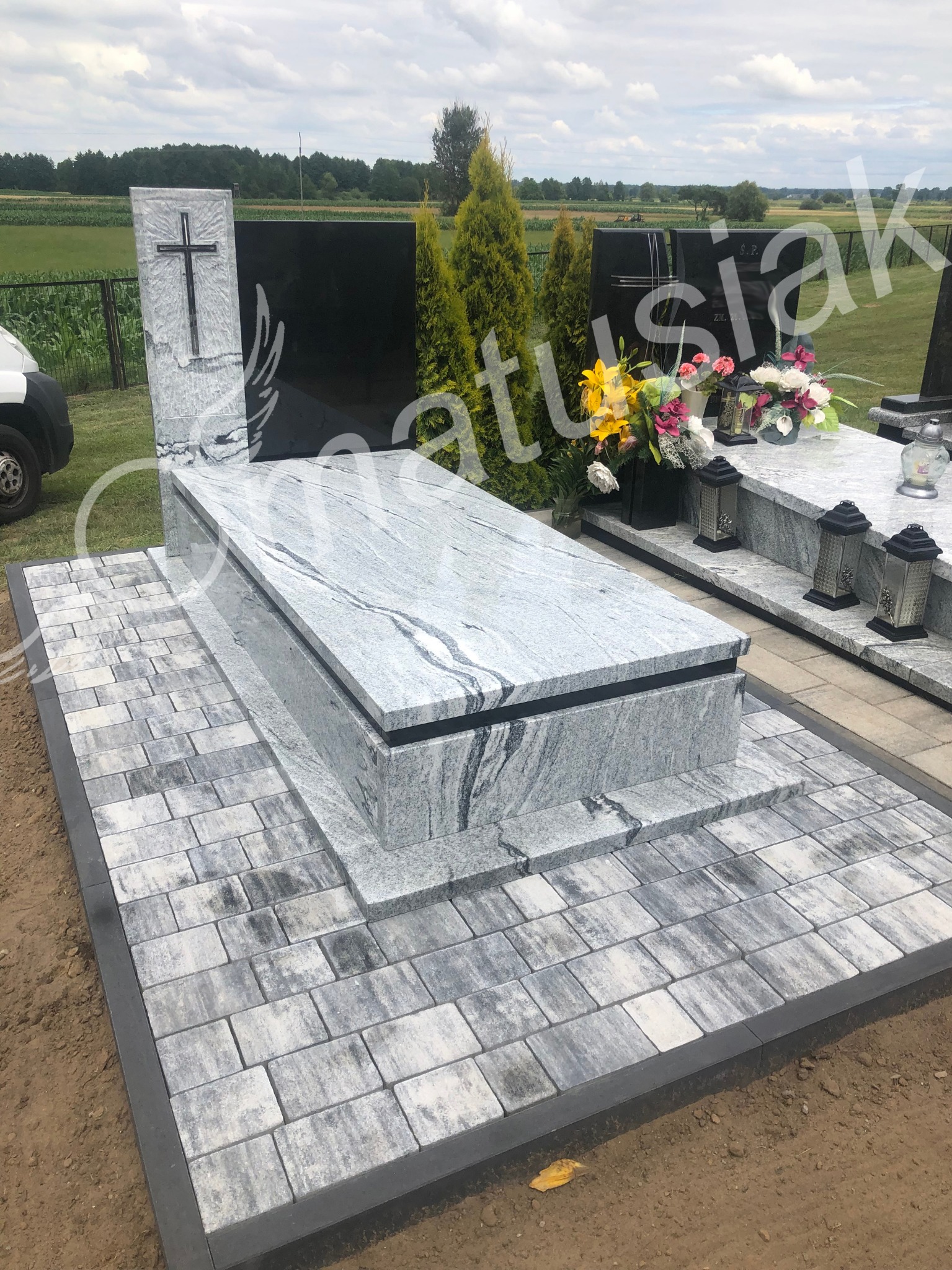 Jasnoszary nagrobek z granitu z krzyżem, otoczony szarą kostką brukową na cmentarzu, w tle zieleń.