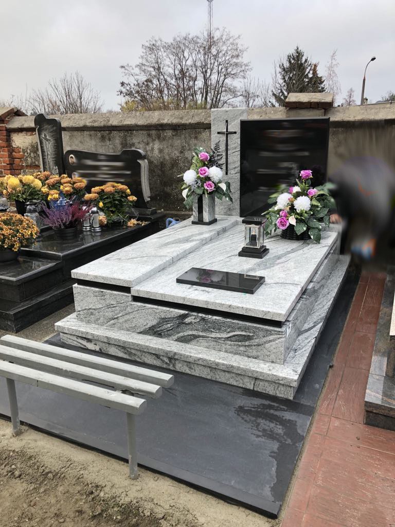 Granitowy nagrobek z jasnoszarego kamienia z ciemnym krzyżem, ozdobiony kwiatami w wazonach i lampionem, ustawiony na cmentarzu z betonowym murem w tle.