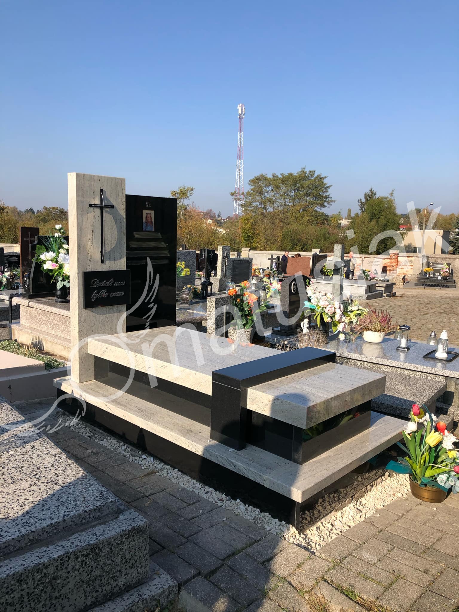 Nowoczesny pomnik z jasnego i czarnego granitu na cmentarzu, z wygrawerowanym krzyżem i sentencją. W tle wieża telekomunikacyjna i inne groby.